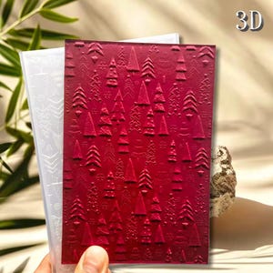 May include: A rectangular 3D embossing folder featuring a deep red surface with a repeating embossed pattern of various stylized Christmas trees and small snowflakes. The folder is translucent white and shows the texture clearly on the red side.