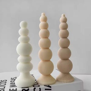 Könnte beinhalten: Drei dekorative Säulenkerzen in verschiedenen Weiß- und Beigetönen. Die Kerzen haben ein einzigartiges Design aus gestapelten Kugeln. Eine Kerze ist glatt, eine gerippt und die dritte ist einfarbig. Die Kerzen stehen auf einer weißen Oberfläche.