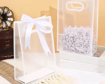 Könnte beinhalten: Zwei transparente, rechteckige Geschenktüten mit weißem Rand. Eine Tüte ist mit einer weißen Satinschleife gebunden. Die andere Tüte enthält geschreddertes Papier. Beide Tüten haben einen Griffausschnitt. Die Tüten stehen auf einer weißen Oberfläche.