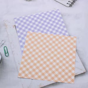 May include: Two square sheets of patterned paper. One is light purple and the other is peach, both with a white gingham pattern. The papers are layered on a white surface with a paper clip and other stationery items.