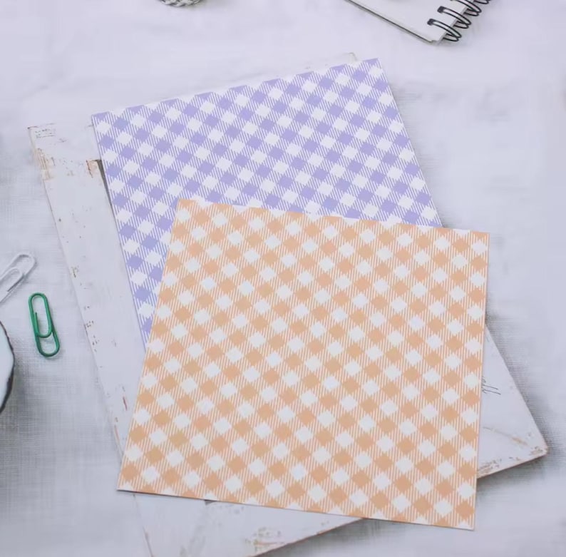 May include: Two square sheets of patterned paper. One is light purple and the other is peach, both with a white gingham pattern. The papers are layered on a white surface with a paper clip and other stationery items.