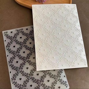 May include: Two rectangular embossing folders featuring a repeating diamond lattice pattern composed of delicate floral vines and small heart shapes. One folder is translucent with dark gray details, while the other is opaque white with raised textures.