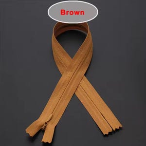 Puede incluir: Cremallera marr&oacute;n con tirador a juego, dispuesta en un patr&oacute;n decorativo. La palabra "Brown" se muestra en un &oacute;valo blanco encima de la cremallera. Los dientes de la cremallera son visibles.