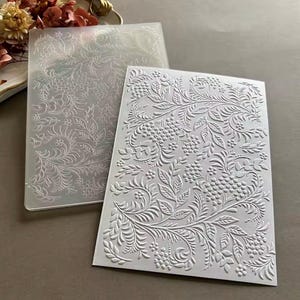 May include: Two rectangular plastic embossing folders featuring a detailed raised pattern of swirling leaves, vines, and clusters of small berries. The folders are translucent and white, designed to create textured impressions on paper or cardstock.