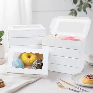 Könnte beinhalten: Weiße Bäckereikartons mit transparenten Fenstern, einige geöffnet, um bunte Donuts zu zeigen. Die Donuts haben verschiedene Beläge und Glasuren, darunter Streusel und Schokolade. Die Kartons sind auf einer weißen Oberfläche gestapelt.