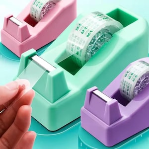 May include: Three colorful tape dispensers in pink, green, and purple. Each dispenser holds a roll of clear tape and has a silver metal blade for cutting. A hand is pulling a piece of tape from the green dispenser.