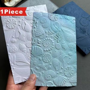 May include: Embossed paper sheets with seashell designs in white, light blue, and dark blue. The sheets feature various shell types, starfish, and coral, creating a nautical theme. The text "1 Piece" is visible on a red banner.