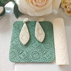 May include: Cream-colored, teardrop-shaped earrings with intricate embossed designs rest on a textured, square, seafoam green surface. A matching embossed roller is positioned to the right. The earrings and roller feature a floral and geometric pattern.