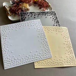 May include: Three square sheets of paper featuring a border of embossed heart shapes. One sheet is white, one is pale yellow, and one is translucent with black printed hearts. The hearts are densely packed at the edges and fade toward the center.