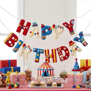 Peut inclure: Une banderole Happy Birthday en feutrine avec des lettres sur le thème du cirque en rouge, bleu et crème, décorées de ballons, tickets et étoiles. En dessous, un gâteau chapiteau, des pots de pop-corn, des bonbonnières et des cadeaux.