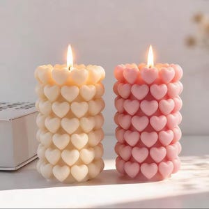 Puede incluir: Dos velas encendidas con forma de corazón. Una es de color amarillo pálido y la otra rosa. Las velas son cilíndricas, con filas de corazones que forman sus cuerpos. Las llamas son brillantes y el fondo es blanco suave. Las velas miden unos 10 cm de alto.