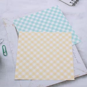 May include: Two square sheets of paper with a gingham pattern. One sheet is yellow and white, and the other is light blue and white. A green paperclip and a white paperclip are visible on the left side of the image.