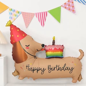 May include: A tan dachshund-shaped foil balloon wearing a red party hat with yellow polka dots. The balloon features a rainbow-coloured cake with a single candle on its back and the text Happy Birthday in black script across its side.