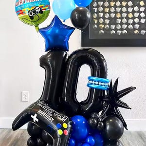 Könnte beinhalten: Eine festliche Ballonanordnung zum zehnten Geburtstag. Enthält schwarze Zahlenballons, einen blauen Stern und verschiedene blaue und schwarze Ballons. Enthält einen Controller-förmigen Ballon mit dem Text "Game On! It's Your Birthday."