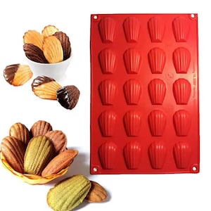 Puede incluir: Un molde de silicona rojo para madeleines con 16 cavidades en forma de concha. Varias madeleines se muestran en un cuenco blanco y una cesta amarilla. Algunas madeleines están bañadas en chocolate. Las madeleines son doradas, marrones claras y verdes.