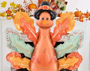May include: A large, orange turkey balloon with leaf-shaped wings in shades of orange and green. The turkey wears a black pilgrim hat with a gold buckle. The balloon is a festive decoration for Thanksgiving or autumn.
