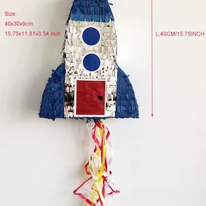 Może przedstawiać: Piñata w kształcie rakiety z niebieskimi frędzlami, srebrnymi metalicznymi panelami, dwoma niebieskimi okrągłymi oknami i czerwonymi kwadratowymi drzwiami. Czerwone, żółte i białe wstążki zwisają u dołu. Wymiary to 40 cm długości, 30 cm szerokości i 9 cm głębokości.