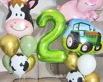May include: A collection of farm-themed balloons, including a cow, pig, and tractor, along with green and gold balloons. A large green number 2 balloon is also present, suggesting a second birthday celebration.