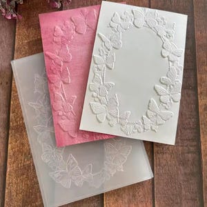 Puede incluir: Tres diseños de papel en relieve con motivos de mariposas. Uno es rosa, otro blanco y el tercero es una plantilla de plástico transparente. Los diseños de mariposas están dispuestos en forma circular, adecuados para manualidades o tarjetas.