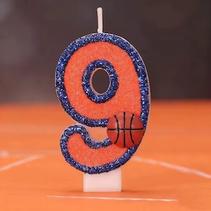 May include: A number 9 birthday candle with an orange body and blue glitter trim. A small basketball is attached to the bottom of the number. The candle has a white base and a white wick.