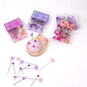 Puede incluir: Un conjunto de alfileres de costura en cajas de plástico transparente y un cojín con forma de gato de madera. Los alfileres tienen cabezas decorativas con forma de gato en rosa y morado. Las cajas están etiquetadas como "Sewing Pins 80pcs".