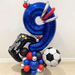 Op de afbeelding: Een feestelijke ballonnenopstelling voor een negende verjaardag. Het bevat een grote blauwe ballon met het cijfer 9, een zwarte videogamecontrollerballon en een voetbalballon. De opstelling bevat rode, zilveren, zwarte en blauwe ballonnen. De controllerballon leest "GAME ON! IT'S YOUR BIRTHDAY".