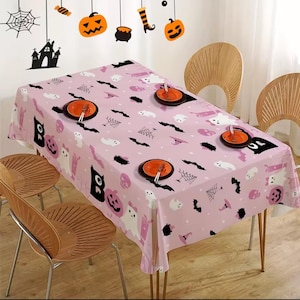Puede incluir: Un mantel rosa con temática de Halloween con un patrón repetido de fantasmas, murciélagos, calabazas y telarañas. La mesa está puesta con platos y cubiertos naranjas. Decoraciones de Halloween cuelgan sobre la mesa.