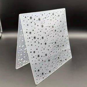 Könnte beinhalten: Ein transparenter, rechteckiger Prägefolder aus Kunststoff mit einem erhabenen, texturierten Muster aus unterschiedlich großen Sternen und kleinen Punkten auf der gesamten Oberfläche. Der Folder ist mittig gefaltet und zeigt das Sternmuster.