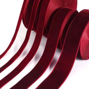 Op de afbeelding: Close-up van een spoel bordeauxrood fluwelen lint. Verschillende strengen van het lint strekken zich uit vanaf de spoel, waarbij de zachte textuur en rijke kleur worden getoond. Het lint is ideaal voor knutselen, cadeauverpakking of elegante projecten.