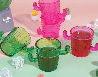 May include: A collection of cactus-shaped drinking glasses in vibrant colors. The glasses are pink, green, and red, with textured surfaces and cactus arm handles. Some glasses contain liquid. Decorative cactus figurines are also present.