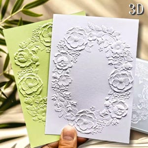 May include: Two rectangular cards featuring a 3D embossed floral wreath design. One card is light green and the other is white. The embossed pattern consists of various flowers and leaves arranged in an oval shape, creating a raised texture.
