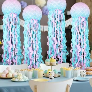 May include: Four hanging jellyfish decorations with blue, pink, and purple streamers. The jellyfish have a round, patterned top and are suspended above a table set for a party. The table is decorated with cupcakes, donuts, and other treats.