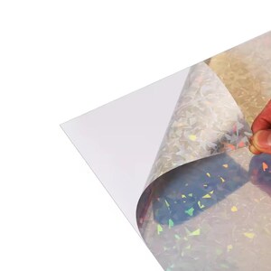 May include: A sheet of iridescent, holographic material with a clear backing is being peeled back, revealing a shimmering, multi-colored surface. The material reflects light in a variety of colors, creating a dazzling effect. The material is likely used for crafts or decorative purposes.