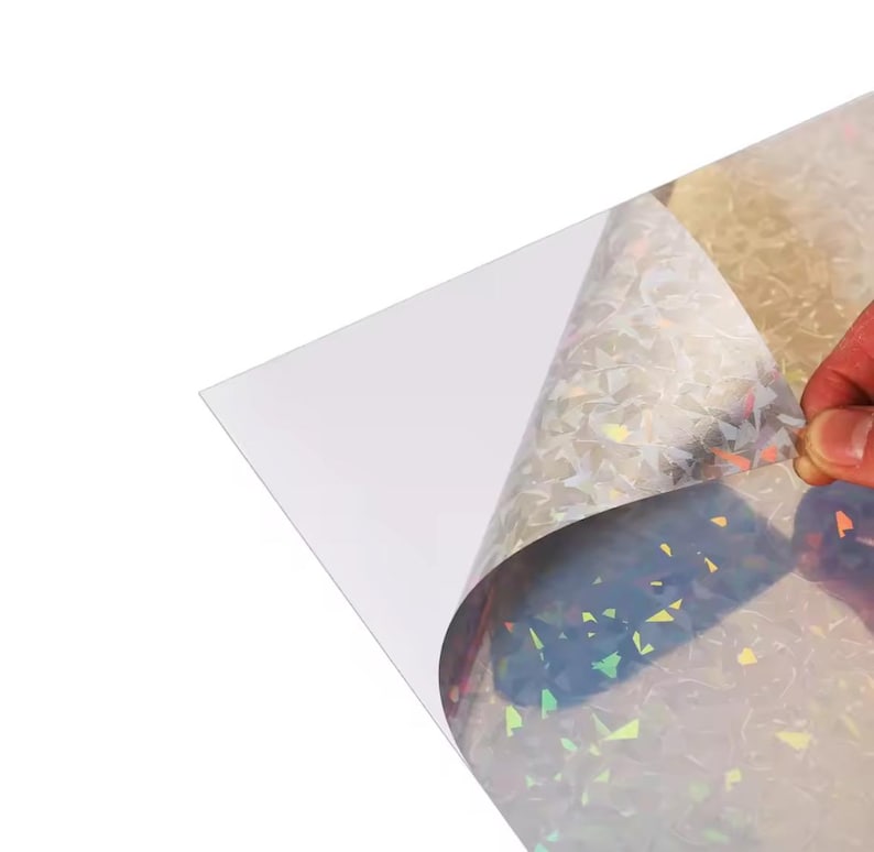 May include: A sheet of iridescent, holographic material with a clear backing is being peeled back, revealing a shimmering, multi-colored surface. The material reflects light in a variety of colors, creating a dazzling effect. The material is likely used for crafts or decorative purposes.