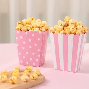 May include: Two pink popcorn containers filled with popcorn. One has white polka dots, the other white stripes. The popcorn is golden brown and looks freshly made. The containers have scalloped edges.