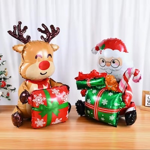 Könnte beinhalten: Zwei Weihnachtsballons: ein Rentier und der Weihnachtsmann. Das Rentier ist braun mit Geweih und roter Nase, hält ein rotes Geschenk mit grünem Band und weißen Schneeflocken. Der Weihnachtsmann trägt einen roten Anzug und Hut und hält ein grünes Geschenk.