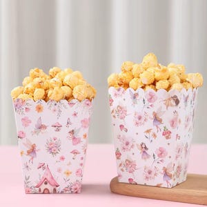Peut inclure: Deux boîtes à popcorn remplies de popcorn. Les boîtes sont blanches avec un motif floral et de fées rose, violet et vert. Le popcorn est de couleur jaune doré. Les boîtes sont posées sur un plateau en bois.