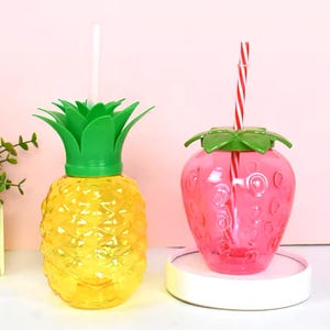 May include: Two plastic cups shaped like a pineapple and a strawberry. The pineapple cup is yellow with a green top and a clear straw. The strawberry cup is pink with a green top and a red and white striped straw.
