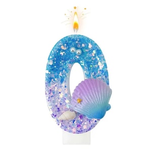 May include: A zero-shaped birthday candle with a mermaid theme. The candle is blue with glitter and a seashell design. The candle is lit with a yellow flame and has a white base.