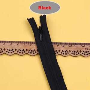 Puede incluir: Una cremallera invisible negra con un tirador negro. La cremallera est&aacute; abierta, revelando los dientes. La palabra "Black" est&aacute; escrita en rojo sobre un &oacute;valo gris encima de la cremallera. Una regla de madera est&aacute; en el fondo.