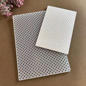 Könnte beinhalten: Ein rechteckiger, durchscheinender Prägefolder aus Kunststoff mit einem sich wiederholenden Muster aus kleinen, gleichmäßig verteilten kreisförmigen Löchern. Ein weißer, rechteckiger Karton mit geprägter Punktstruktur liegt darauf.