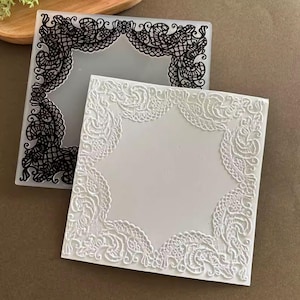 Puede incluir: Dos moldes cuadrados decorativos. Uno es blanco con un diseño de encaje en relieve adornando los bordes. El otro es translúcido con un diseño de encaje negro. Ambos tienen un centro cuadrado y abierto.