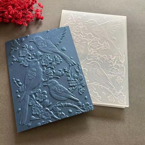 May include: Two embossed cardstock pieces. One is a deep blue with a raised design of birds perched on flowering branches. The other is a clear, translucent piece with the same design. Red floral accents are in the background.