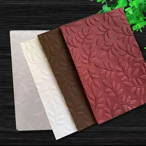 May include: A set of four rectangular embossing folders featuring a repeating leaf pattern. The folders are presented in a gradient of colors, including translucent, white, dark brown, and deep red, each displaying the same embossed foliage design.