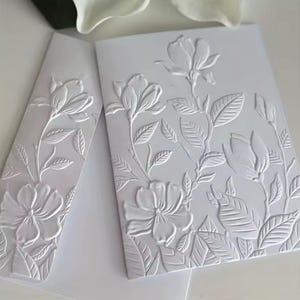 May include: White embossed floral stationery set featuring a detailed magnolia design. The set includes a card and an envelope, both with raised floral patterns. The design includes flowers, leaves, and stems, creating a textured, elegant look.
