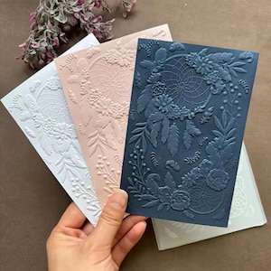 Op de afbeelding: Drie rechthoekige embossing folders in wit, lichtroze en donkerblauw. Elke folder heeft een verhoogd, gestructureerd ontwerp van een dromenvanger omringd door gedetailleerde bloemen- en bladpatronen voor een reliëfeffect.