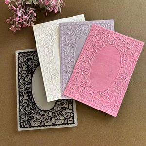 May include: Three rectangular embossing folders in black, white, and pink, each featuring an intricate, raised floral and vine pattern surrounding a smooth, central oval frame. The folders are arranged in a layered, overlapping stack.