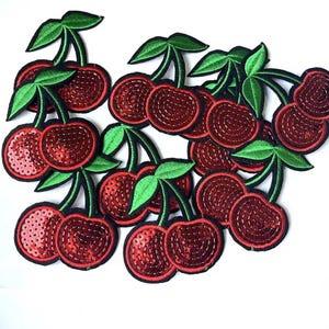 May include: Embroidered cherry patches with red sequins and green leaves. The cherries have a black outline and are arranged on a white background. These patches can be used for clothing or craft projects.