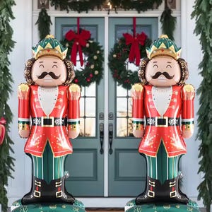 Puede incluir: Dos figuras inflables de cascanueces frente a una puerta decorada para las fiestas. Los cascanueces son rojos, verdes y dorados, con botas negras y una base verde. Tienen bigote y corona.