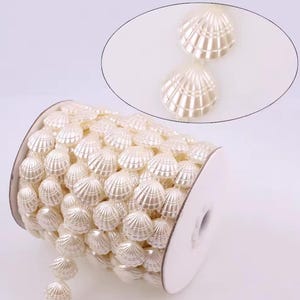 Op de afbeelding: Een rol crèmekleurige schelpvormige kralen. De kralen zijn in een doorlopende lijn gerangschikt, met een close-up van twee afzonderlijke kralen in de rechterbovenhoek. De kralen hebben een parelmoerglans en worden waarschijnlijk gebruikt voor knutselen of versiering.